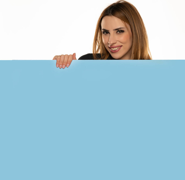 una giovane donna eccitata fa annunci sbircia dietro una tavola blu vuota su uno sfondo bianco - Foto, immagini