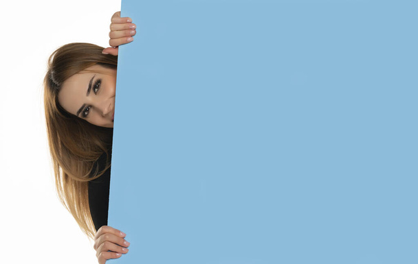 mladá reklamní vzrušená žena nakukuje za prázdné modré prkno na bílém pozadí - Fotografie, Obrázek