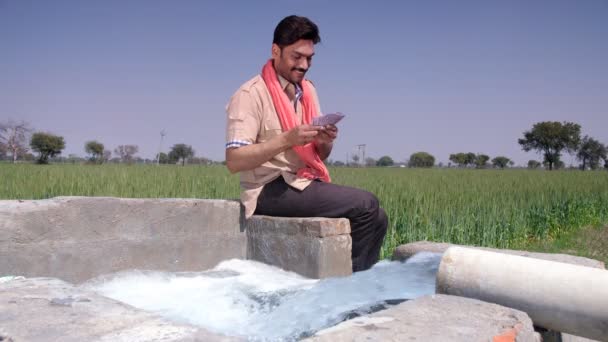 農家の補助金、バンパー作物の収穫、農業利益、毎月の収入-明るいインドの農家は、井戸の近くに座っている間にお金を数える。彼の農業分野で親指アップジェスチャーを行う伝統的なインドの村人 - 映像、動画