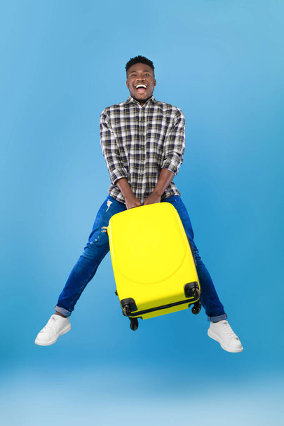 Czas na podróż. Pełna długość podekscytowany młody czarny człowiek skacze z jasną walizką nad niebieskim tle studio - Zdjęcie, obraz