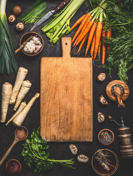 Food background with cutting board and various organic farm vegetables on dark rustic table with kitchen utensils, herbs and spices, top view. Frame. Copy space. Vegan and vegetarian food concept. - Foto, Imagem