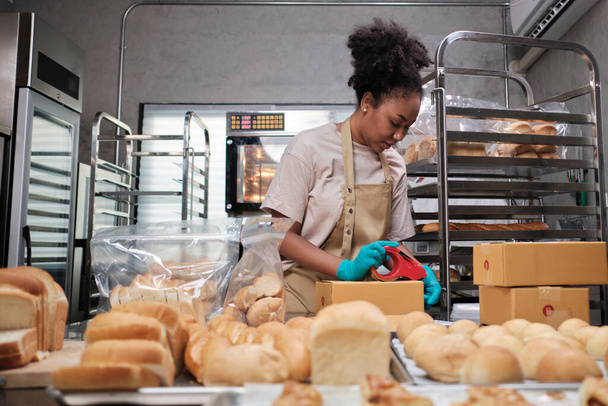 Bakkerij startup kleine bedrijven levering. Een Afro-Amerikaanse vrouwelijke kok is het verpakken van handgemaakte en vers gebakken brood en gebak in dozen en het verzenden voor online aankoop van klanten in de culinaire keuken. - Foto, afbeelding