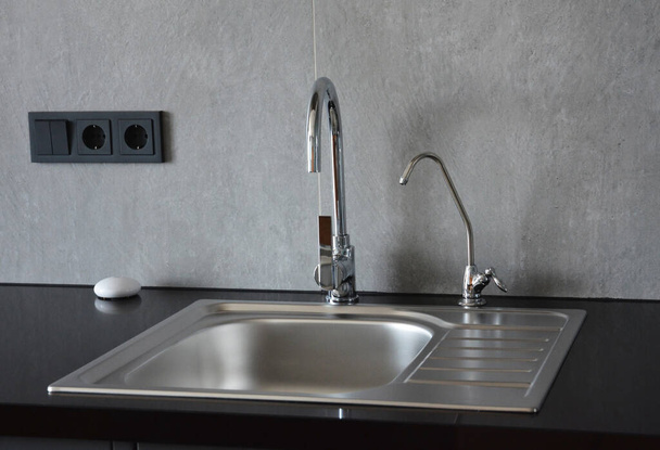 A metal kitchen sink on a black kitchen countertop with two water faucets, where one is a small filter faucet for drinking water. - Foto, Imagen