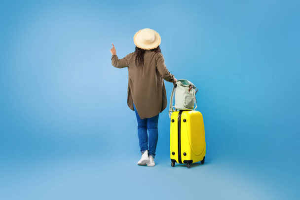 Back view of young black woman with luggage pointing, choosing tourist tour on blue studio background, copy space - Foto, afbeelding
