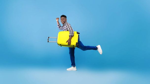 空港に走っているスーツケース付きの興奮した黒い男の完全な長さ,青のスタジオの背景で夏休みを開始する準備ができて - 写真・画像