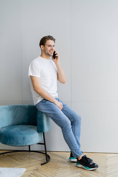 full length view of smiling man in jeans talking on smartphone near armchair and grey wall - Photo, Image