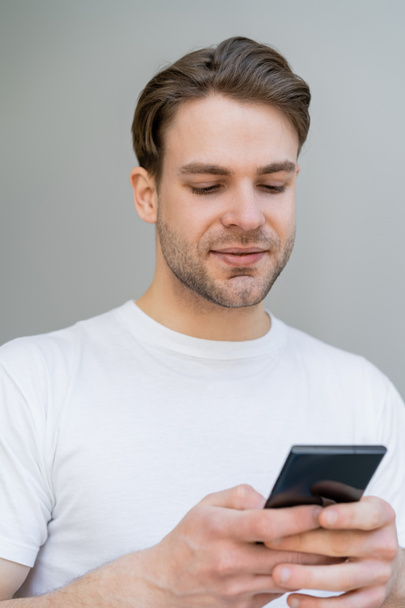 young smiling man in white t-shirt using smartphone isolated on grey - Photo, Image