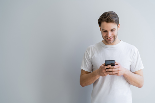cheerful young man in white t-shirt chatting on mobile phone on grey with copy space - Foto, imagen