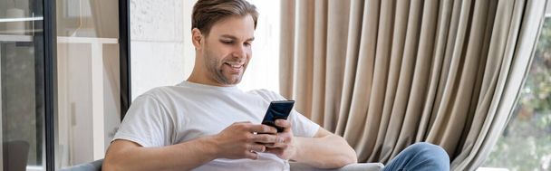 happy man messaging on smartphone while sitting at home near beige curtain, banner - Φωτογραφία, εικόνα