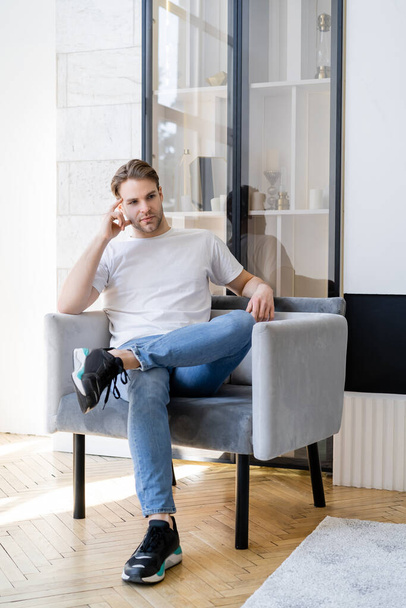 thoughtful man in white t-shirt sitting in armchair with crossed legs and looking away - Foto, Bild