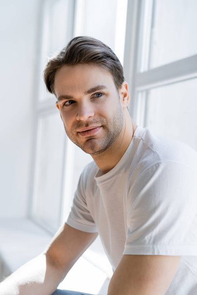 portrait of positive man in white t-shirt looking away near window at home - Fotografie, Obrázek