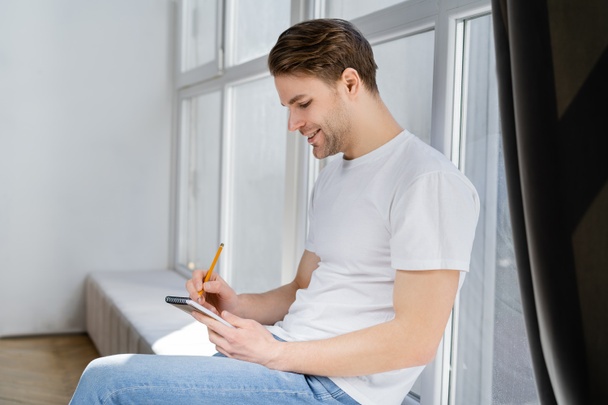 side view of pleased man sitting on windowsill and writing in notebook - Photo, Image