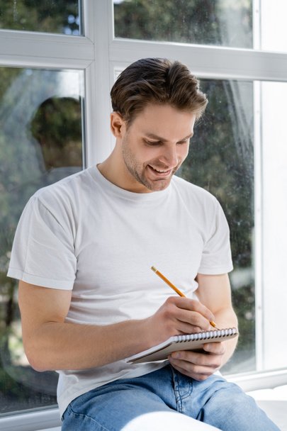homem sorridente escrevendo no diário perto da janela em casa - Foto, Imagem