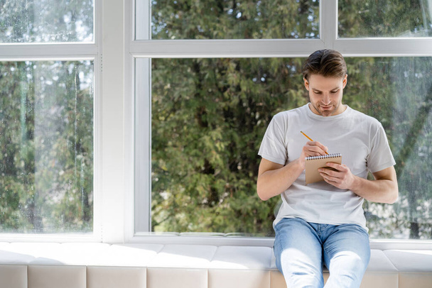 young man in white t-shirt and jeans sitting on windowsill and writing in diary - Photo, Image