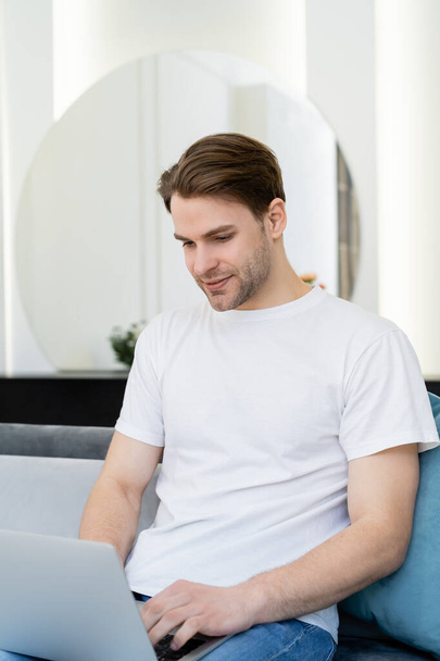 young and smiling man in white t-shirt working on laptop at home - Foto, Bild