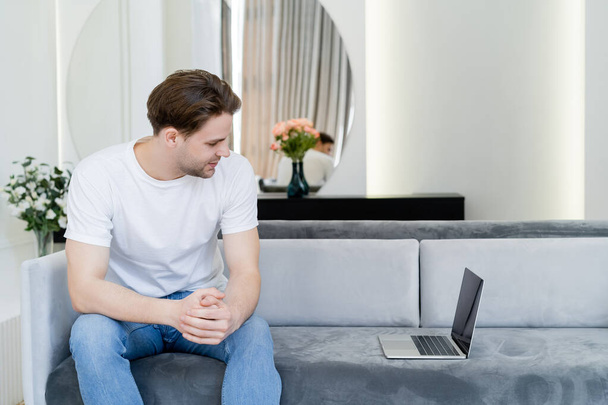 young man sitting on sofa in living room and looking at laptop with blank screen - Фото, изображение