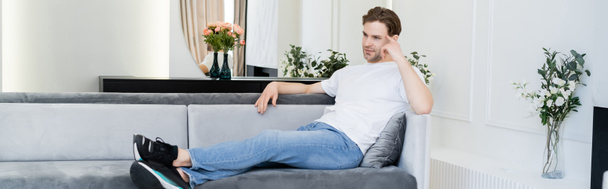 vista completa del hombre reflexivo sentado en el sofá en la sala de estar con decoración floral, pancarta - Foto, imagen
