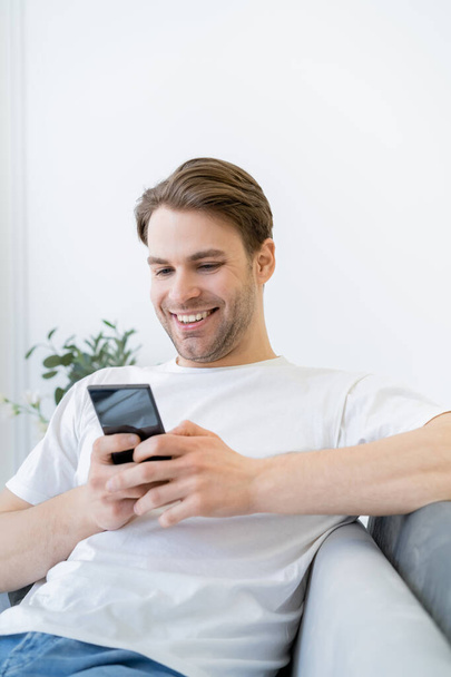 cheerful young man in white t-shirt messaging on cellphone while sitting at home - Фото, изображение