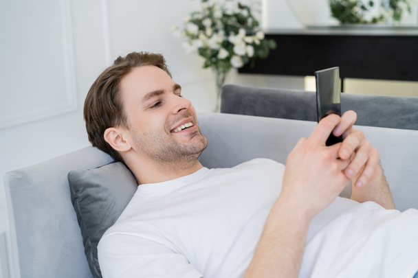 pleased young man resting on couch and chatting on smartphone - Foto, afbeelding