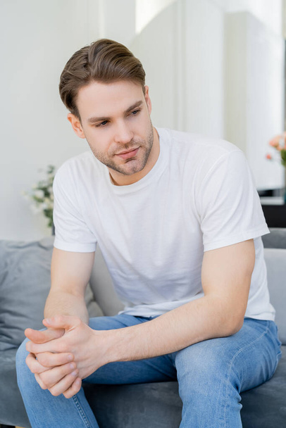 young man in white t-shirt sitting with clenched hands in living room - Foto, imagen