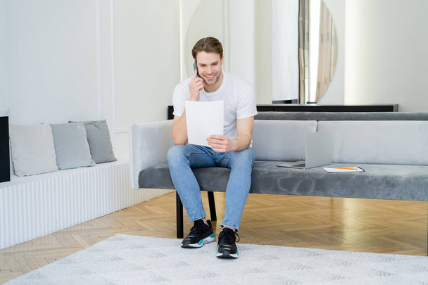 full length view of man with paper talking on mobile phone near laptop on sofa - Φωτογραφία, εικόνα