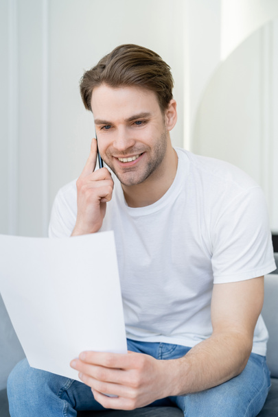 hombre sonriente en camiseta blanca mirando el papel mientras habla por teléfono celular - Foto, imagen