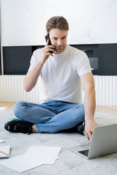 jeune homme assis sur le sol avec les jambes croisées et parlant sur smartphone près d'un ordinateur portable - Photo, image