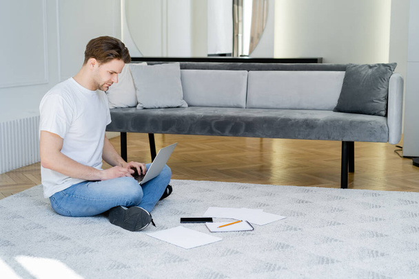 full length of man sitting on floor in living room and typing on laptop - Photo, Image