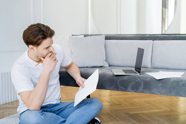 thoughtful man with pencil and paper sitting with crossed legs near laptop with blank screen - Foto, Imagem
