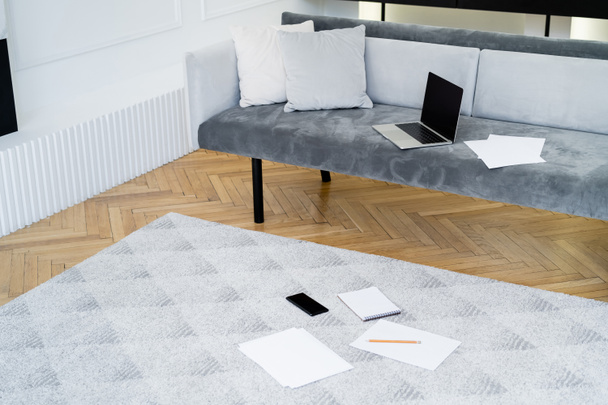 ноутбук, смартфон и пустые бумаги на диване и полу в гостиной - Фото, изображение
