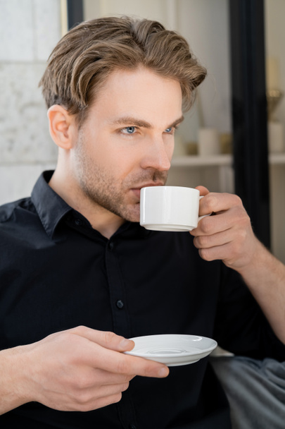 férfi fekete ingben tartó csészealj és iszik kávét fehér csésze - Fotó, kép