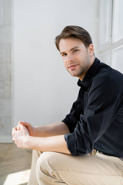 young and stylish man sitting with clenched hands and looking at camera - Foto, immagini