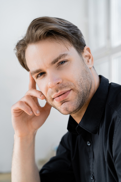portrait of young man in black shirt holding hand near face while looking at camera - Photo, Image