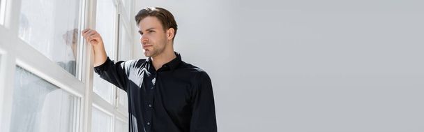 pensive man in black shirt standing near window and looking away, banner - Фото, изображение