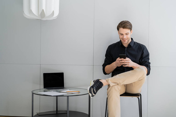 happy man typing on smartphone while sitting with crossed legs near laptop with blank screen - Photo, Image