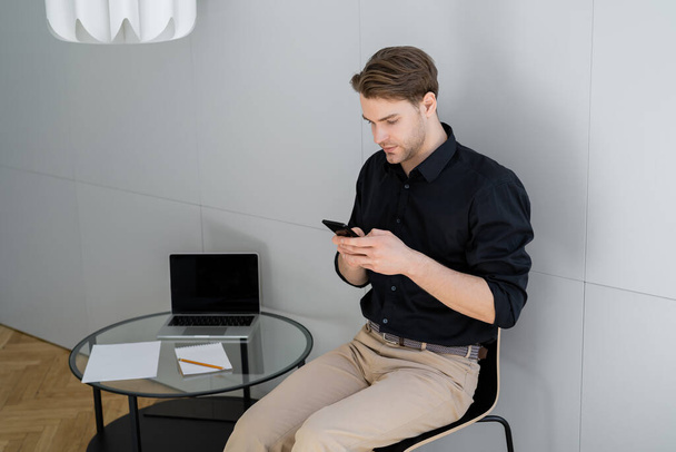 man sitting near laptop with blank screen on coffee table and messaging on smartphone - Foto, afbeelding