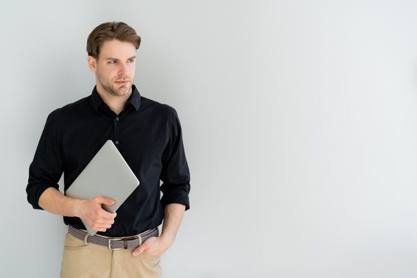 man in black shirt holding laptop while standing with hand in pocket isolated on grey - Photo, Image