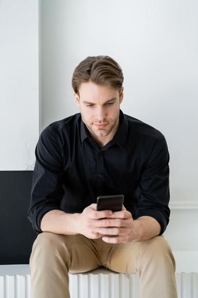 stylish man in black shirt sitting at home and messaging on smartphone - Foto, imagen
