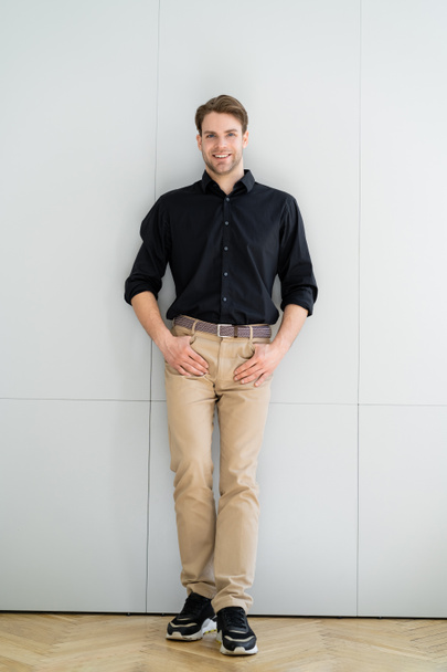 vista completa del hombre sonriente en camisa negra de pie con pulgares en los bolsillos  - Foto, Imagen