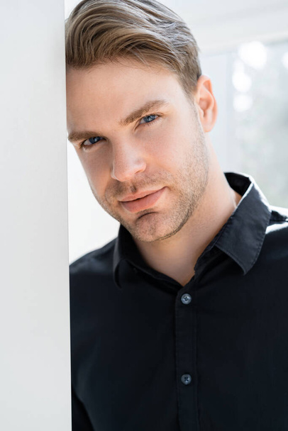 portrait of young man in black shirt looking at camera while leaning on wall - Fotografie, Obrázek