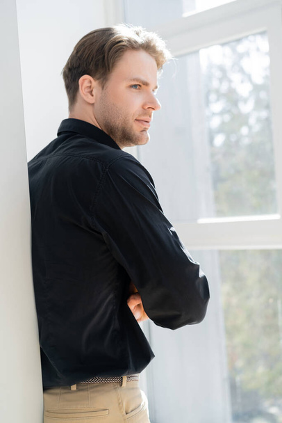 junger Mann in schwarzem Hemd steht am Fenster und schaut weg - Foto, Bild
