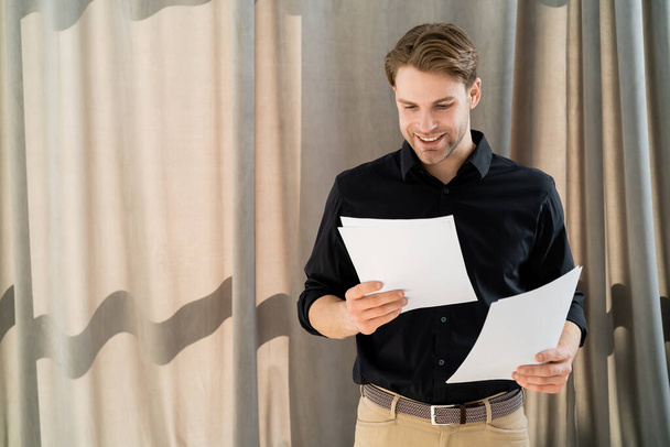 young and positive man in black shirt looking at papers near beige curtain - 写真・画像