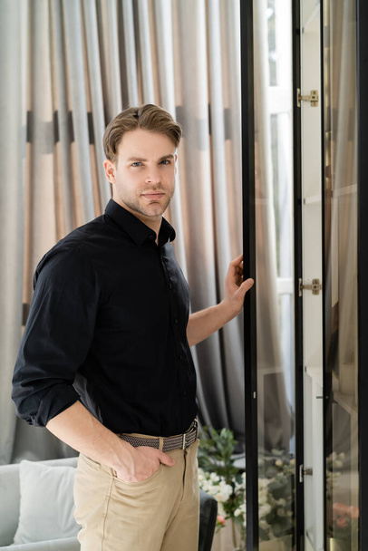 man in black shirt standing with hand in pocket near window and beige curtain at home - Foto, Bild