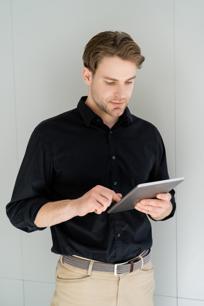 man in black t-shirt using digital tablet while standing near grey wall - Фото, изображение