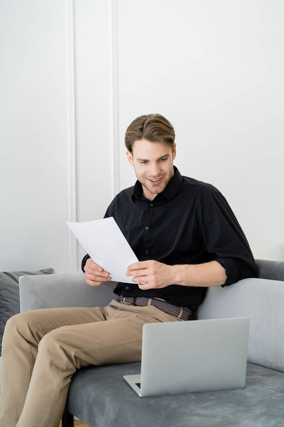 smiling man with document sitting on couch and looking at laptop - Foto, Bild