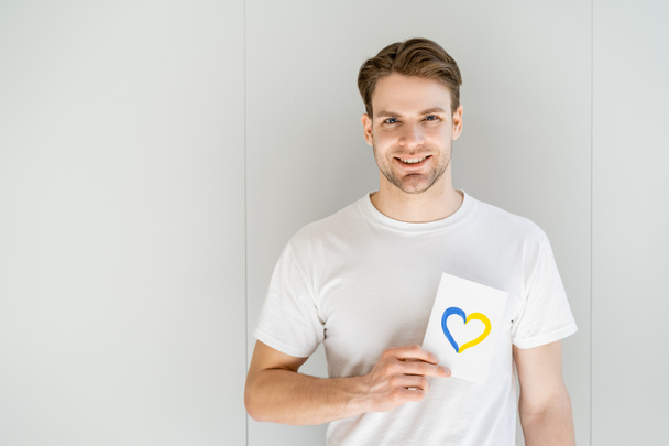 happy man holding white card with blue and yellow heart on grey background - Photo, Image
