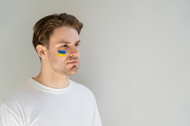 young patriotic man with ukrainian flag painted on face looking away isolated on grey - Foto, immagini
