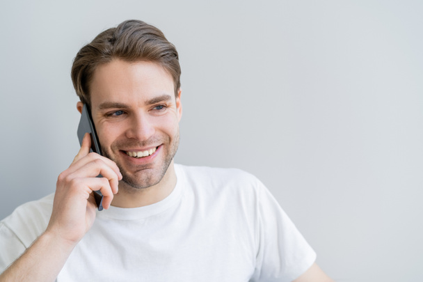 happy young man in white t-shirt talking on smartphone isolated on grey - Photo, Image
