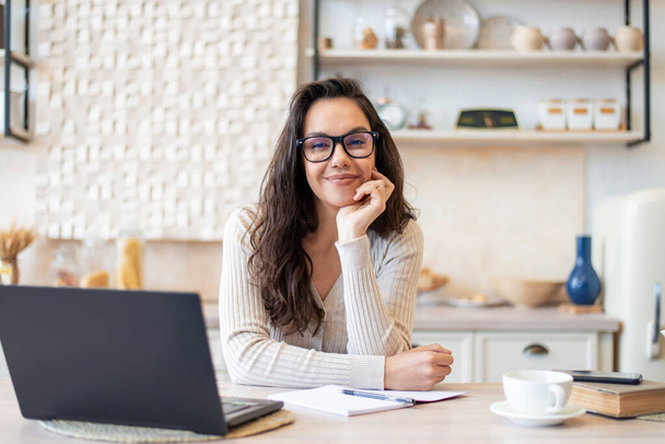 Mujer feliz en gafas y ropa casual mirando y sonriendo a la cámara, sentado con el ordenador portátil en la mesa en el interior de la cocina - Foto, Imagen