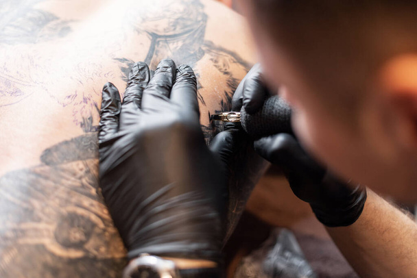 Zbliżenie męskich dłoni w czarnych rękawiczkach z maszyną do tatuażu robi tatuaż atramentem. Tatuaż artysta w pracy - Zdjęcie, obraz